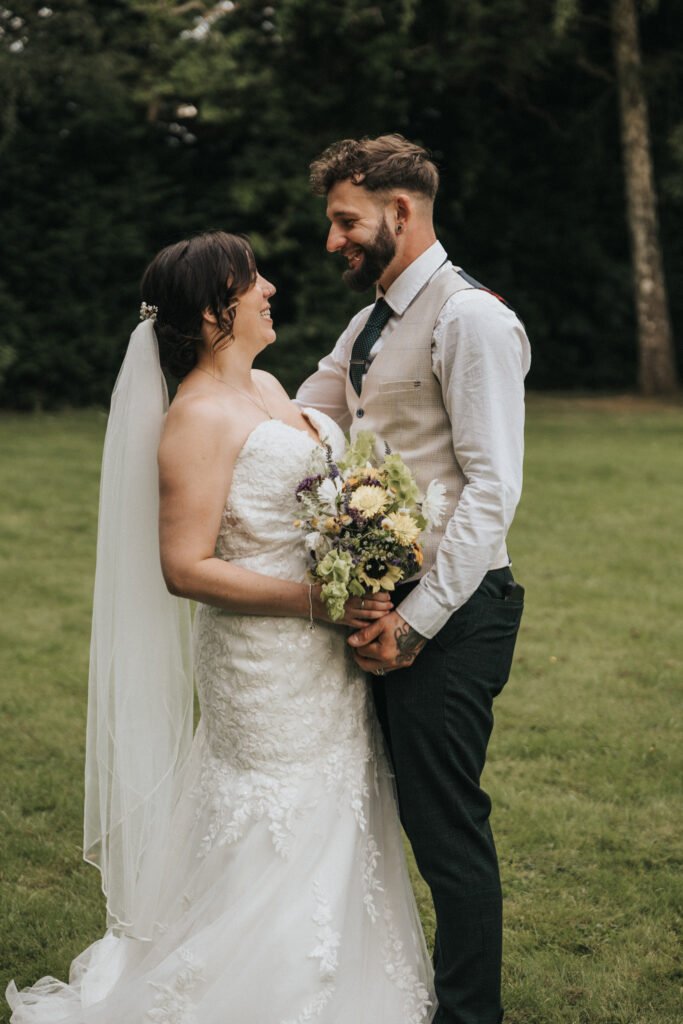 A bride in a strapless white gown and veil smiles at her groom as their wedding photographer captures the moment. They stand closely, gazing at each other, on green grass with trees in the background. © Aimee Lince Photography, Lincolnshire photographer