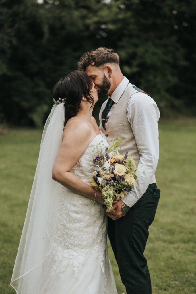 A bride in a white lace gown and veil holds a bouquet whilst sharing an intimate moment with her groom, expertly captured by a wedding photographer. They stand close on green grass with leafy trees in the background. © Aimee Lince Photography, Lincolnshire photographer