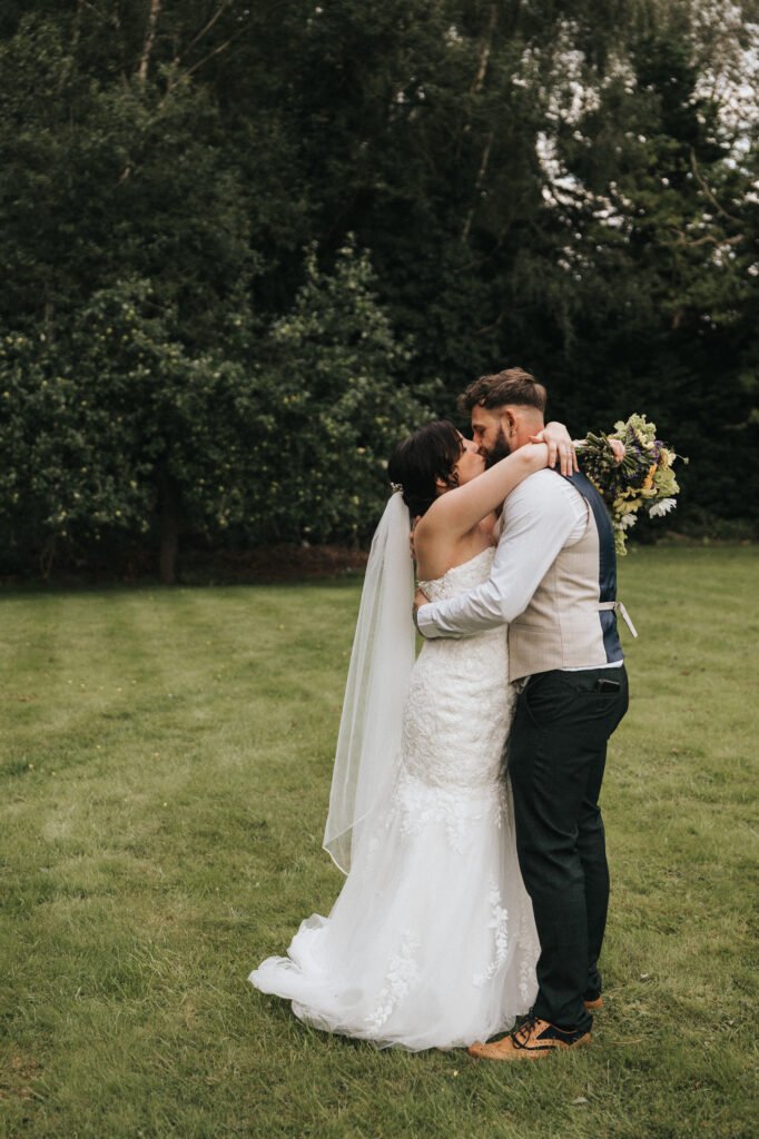 A bride in a white lace gown and veil embraces a groom on a grassy lawn as their wedding photographer captures the tender moment. The bride holds a bouquet behind his back as they stand close, touching foreheads, framed by trees. © Aimee Lince Photography, Lincolnshire photographer