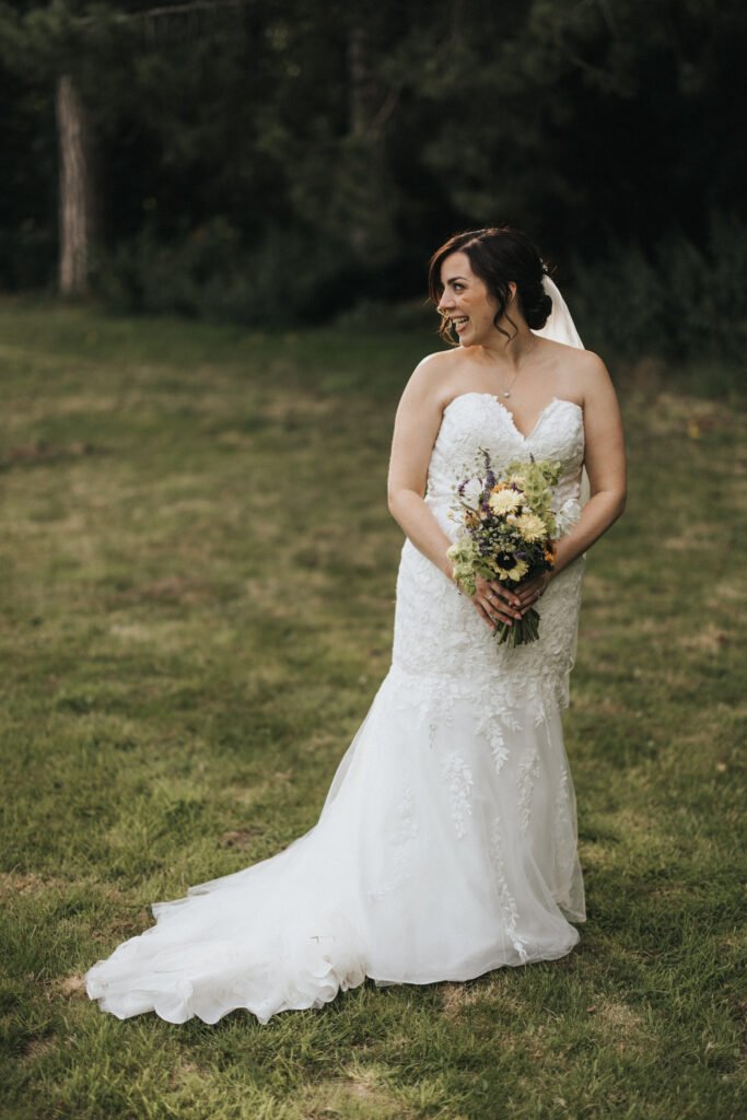 A bride stands outdoors on grass, wearing a strapless white lace wedding dress with a train and veil. She holds a bouquet, smiles, and looks to her left as a wedding photographer captures her amidst lush greenery and trees in the background. © Aimee Lince Photography, Lincolnshire photographer