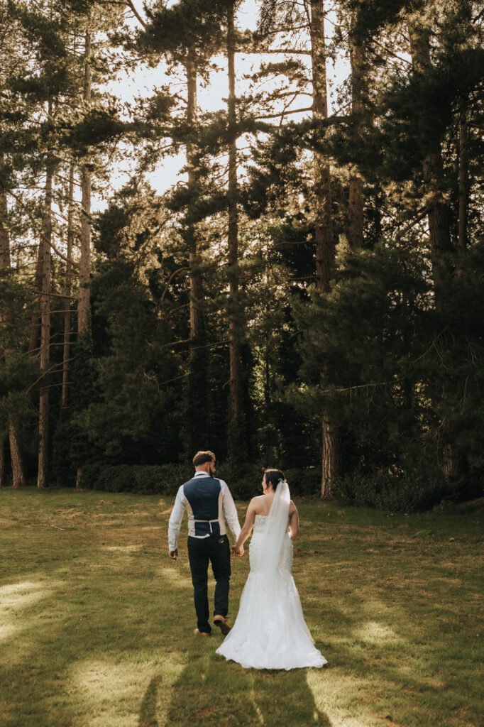 A bride in a white dress and veil walks hand in hand with a groom across a grassy field, sunlight streaming through tall pines. Captured perfectly by their wedding photographer, their backs are facing the camera. © Aimee Lince Photography, Lincolnshire photographer