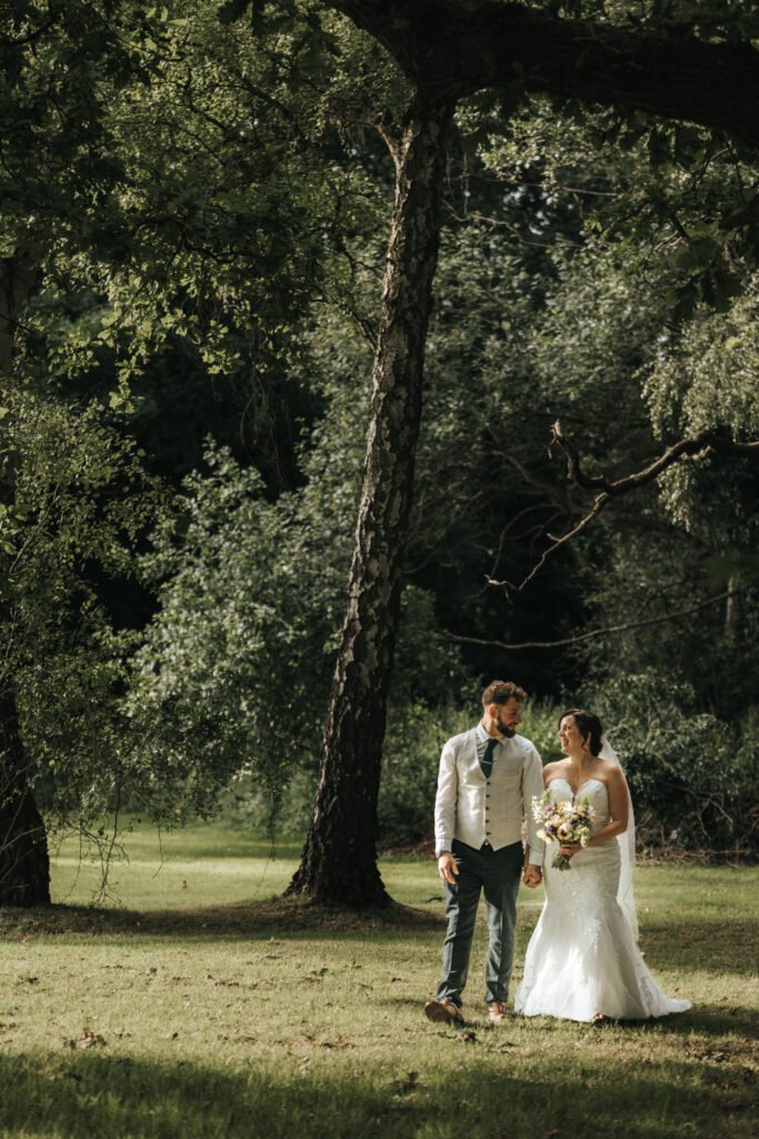 A bride in a white dress and a groom in a waistcoat and tie walk hand in hand on sunlit grass, smiling at each other as their wedding photographer captures the moment, surrounded by tall trees and lush greenery in a peaceful outdoor setting. © Aimee Lince Photography, Lincolnshire photographer