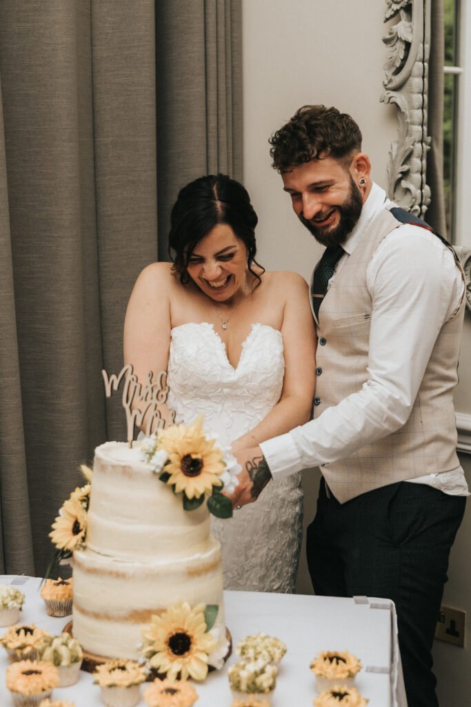 A smiling bride in a strapless white lace dress and a groom in a beige waistcoat and tie cut a two-tier sunflower wedding cake, as their photographer captures the joyful scene in a bright, elegant room filled with cupcakes and sunflower decorations. © Aimee Lince Photography, Lincolnshire photographer