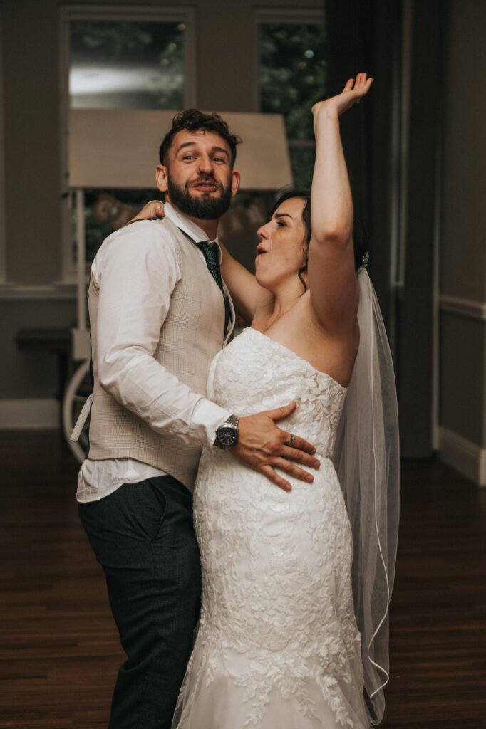 A bride in a white lace strapless dress and veil dances joyfully with her groom as their wedding photographer captures the moment. Her arms are raised, eyes closed, while he smiles and holds her waist on the wooden floor indoors. © Aimee Lince Photography, Lincolnshire photographer
