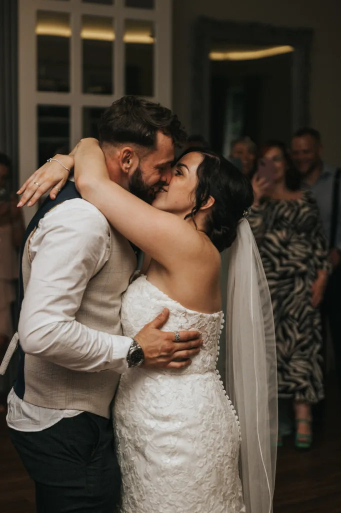A bride in a white lace wedding dress and veil embraces and kisses a groom in a white shirt and waistcoat on the dance floor, while guests smile in the softly lit background—captured beautifully by the wedding photographer. © Aimee Lince Photography, Lincolnshire photographer
