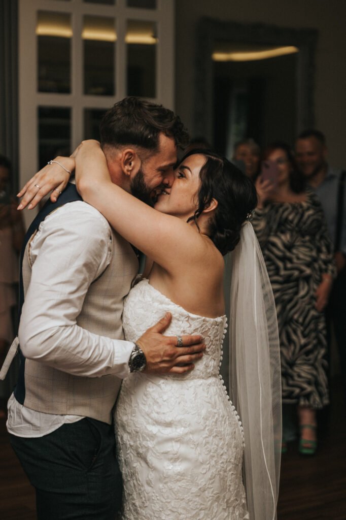 A bride in a white lace wedding dress and veil embraces and kisses a groom in a white shirt and waistcoat on the dance floor, while guests smile in the softly lit background—captured beautifully by the wedding photographer. © Aimee Lince Photography, Lincolnshire photographer