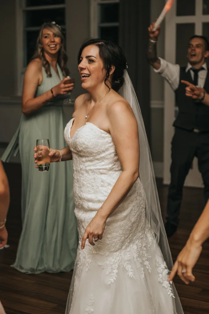 A bride in a strapless white lace gown and veil smiles whilst holding a drink, dancing at an indoor wedding reception as a wedding photographer captures the joy. A bridesmaid in mint green and a man in a waistcoat smile behind her, enjoying the celebration. © Aimee Lince Photography, Lincolnshire photographer