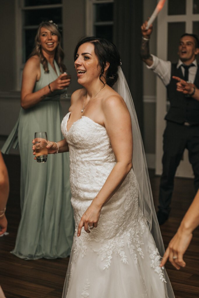 A bride in a strapless white lace gown and veil smiles whilst holding a drink, dancing at an indoor wedding reception as a wedding photographer captures the joy. A bridesmaid in mint green and a man in a waistcoat smile behind her, enjoying the celebration. © Aimee Lince Photography, Lincolnshire photographer
