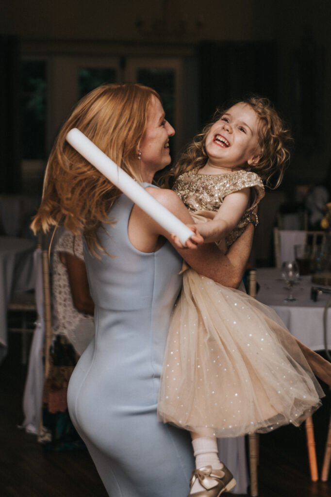 A woman in a pale blue dress spins and holds a smiling young girl in a sparkly gold dress. The child laughs joyfully, her curly hair bouncing, as a wedding photographer captures this heartwarming moment indoors among round tables and chairs. © Aimee Lince Photography, Lincolnshire photographer