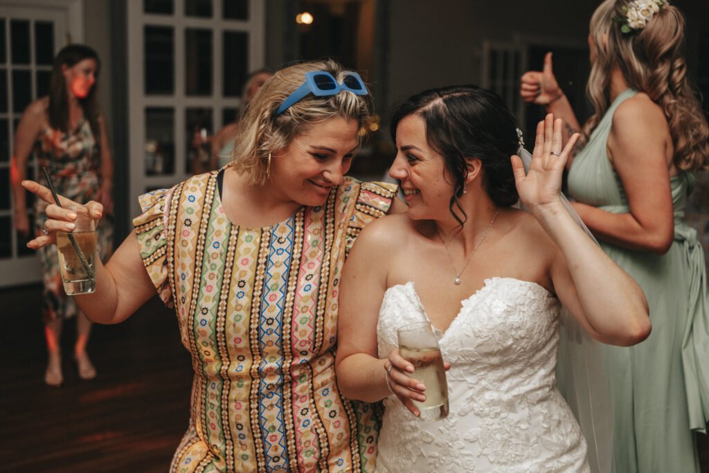 Two women laugh at a party. One, in a patterned dress and blue sunglasses, holds a drink. The other wears a white strapless wedding dress, raising her hand. Captured by a wedding photographer, the scene glows with soft lights and happy guests in the background. © Aimee Lince Photography, Lincolnshire photographer