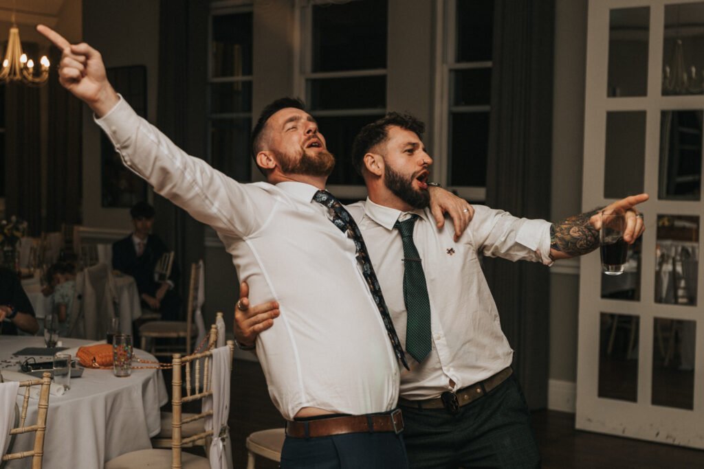 Two men in dress shirts and ties joyfully sing with arms outstretched at an indoor event, captured candidly by a wedding photographer. One holds a drink and embraces the other, whilst tables and chairs fill the softly lit background. © Aimee Lince Photography, Lincolnshire photographer