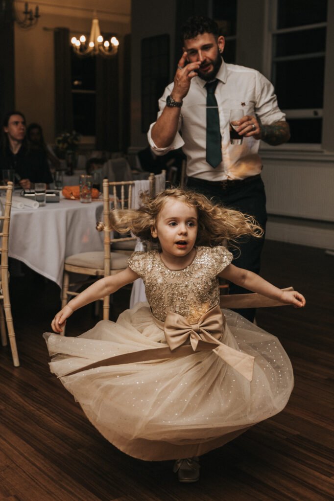 A young girl in a sparkly, gold dress twirls on a wooden floor as a wedding photographer captures the moment. Behind her, a bearded man in a white shirt and tie holds a drink, gesturing whilst guests sit at decorated tables in the warmly lit room. © Aimee Lince Photography, Lincolnshire photographer