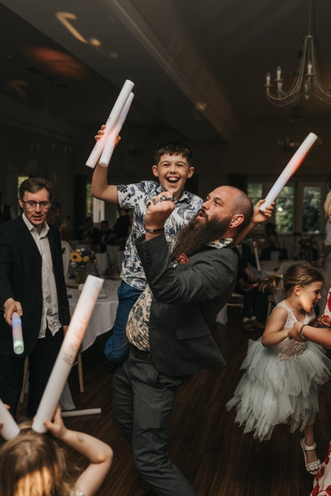 A bearded man in a suit joyfully lifts a smiling boy holding a glowing stick at a lively indoor event. A wedding photographer captures partygoers, including children and adults, dancing with light sticks under chandeliers in the warmly lit room. © Aimee Lince Photography, Lincolnshire photographer