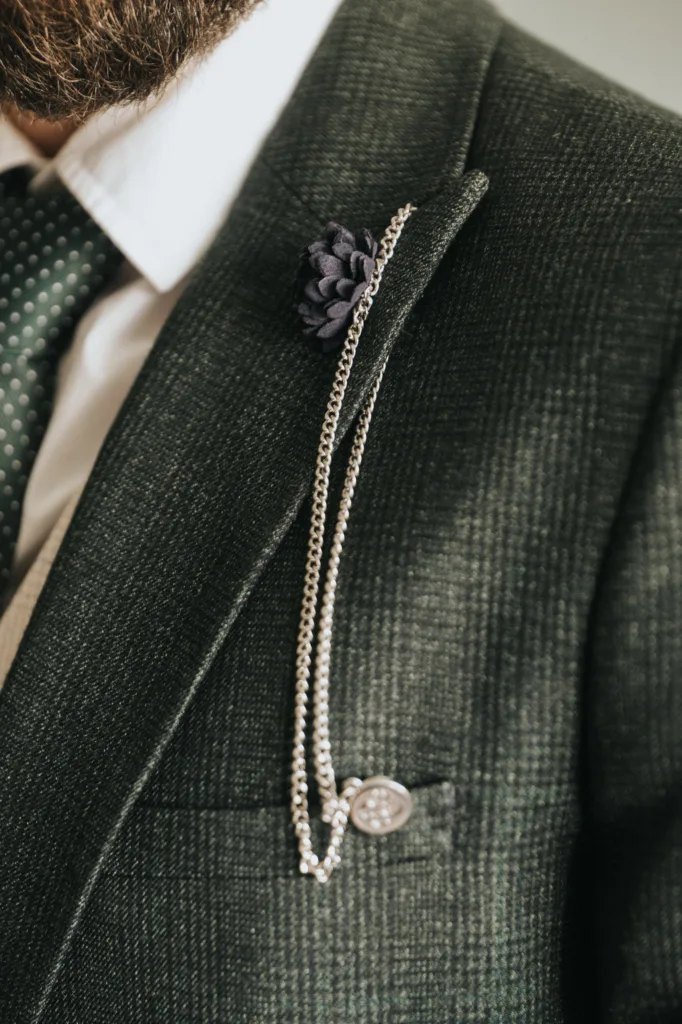 Close-up of a man's green textured suit jacket with a white shirt and green polka-dot tie, captured by a wedding photographer. A silver chain with a round pin is on the lapel, along with a dark purple flower buttonhole. Part of a bearded face shows. © Aimee Lince Photography, Lincolnshire photographer