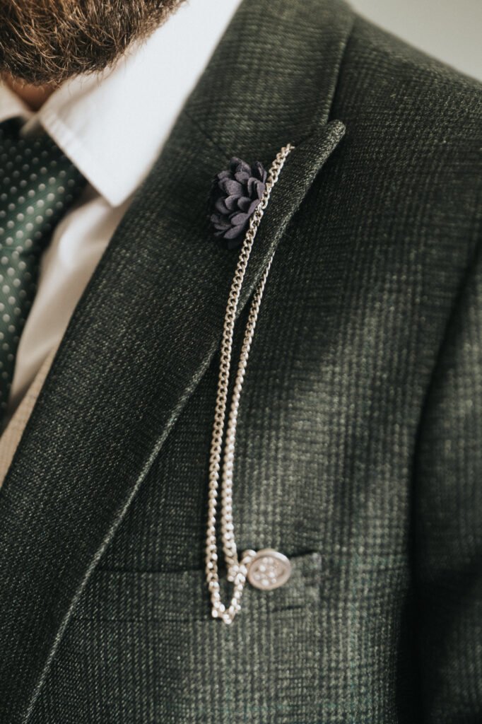 Close-up of a man's green textured suit jacket with a white shirt and green polka-dot tie, captured by a wedding photographer. A silver chain with a round pin is on the lapel, along with a dark purple flower buttonhole. Part of a bearded face shows. © Aimee Lince Photography, Lincolnshire photographer
