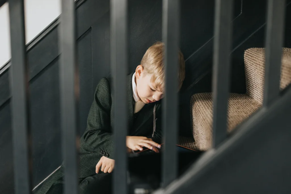 A young boy in a dark green suit sits on the floor by carpeted stairs, focused on a tablet. Captured by a wedding photographer, he is framed by black stair railings and soft lighting illuminating his face and hair. © Aimee Lince Photography, Lincolnshire photographer