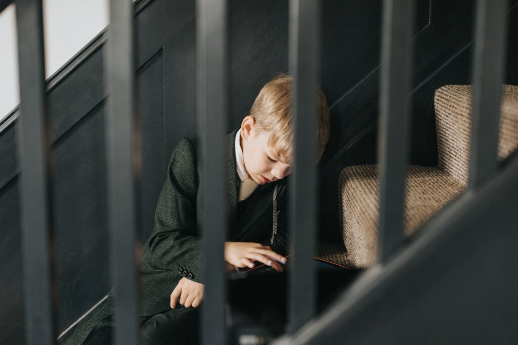 A young boy in a dark green suit sits on the floor by carpeted stairs, focused on a tablet. Captured by a wedding photographer, he is framed by black stair railings and soft lighting illuminating his face and hair. © Aimee Lince Photography, Lincolnshire photographer