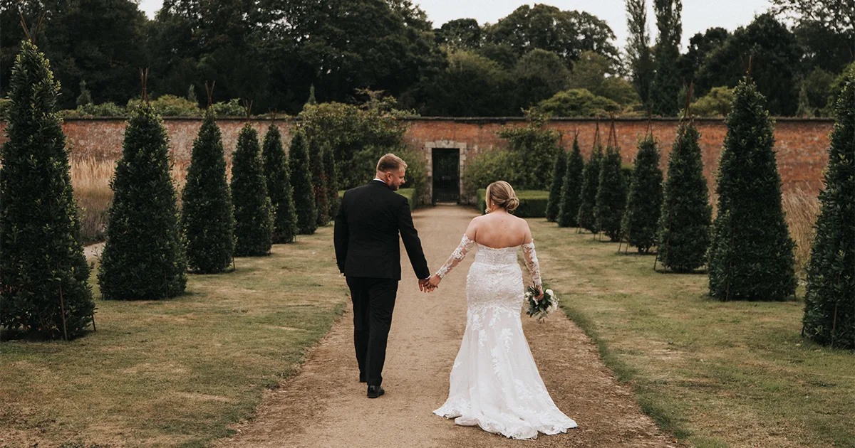 A bride in a white lace gown and a groom in a black suit walk hand in hand down a garden path at Elsham Hall, flanked by manicured topiary trees. Captured from behind, this moment is perfect for any wedding photographer’s portfolio. © Aimee Lince Photography, Lincolnshire photographer