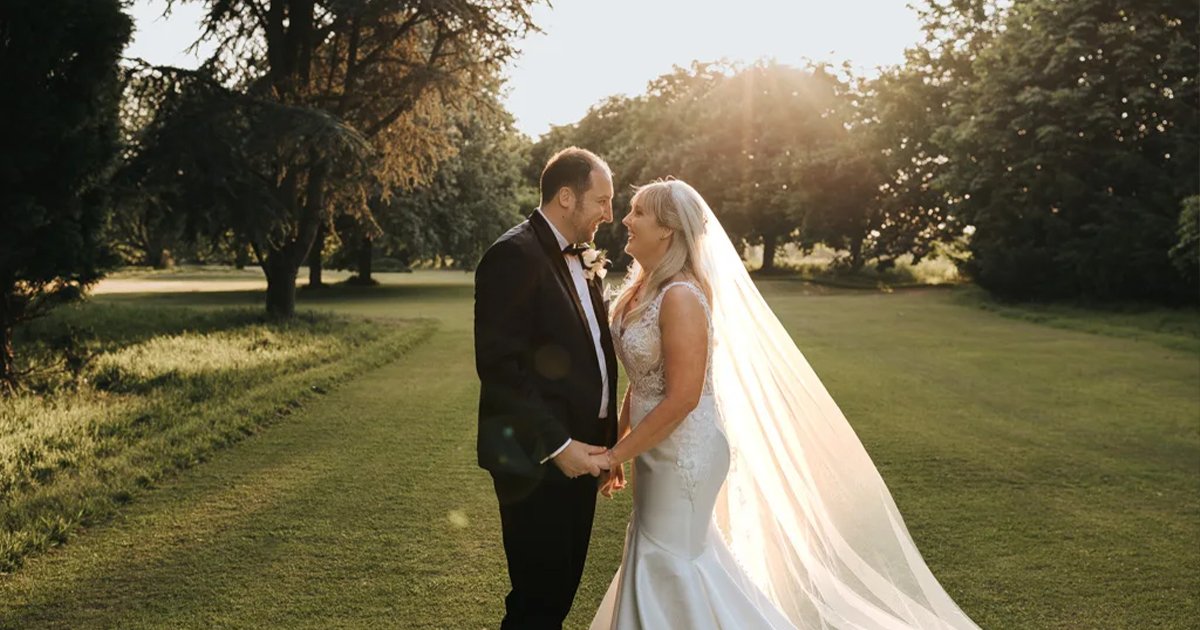 Gemma & Nick stand facing each other on a grassy lawn at sunset, holding hands. The bride’s white dress and long veil glow in the fading light as sunlight filters through trees at their Summer Wedding at Wootton Hall Estate. © Aimee Lince Photography, wedding and lifestyle photographer in Lincolnshire