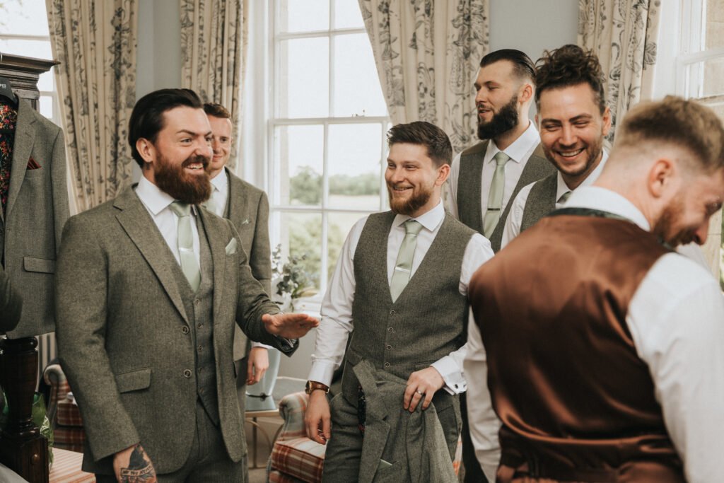 A group of men in grey suits and light green ties laugh and talk together in a bright room with large windows and patterned curtains. One man in the foreground wears a brown waistcoat with his back to the camera. The mood is cheerful and relaxed. © Aimee Lince Photography, Lincolnshire photographer