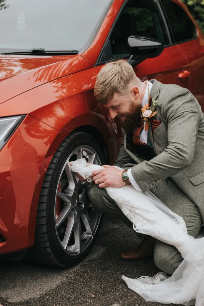 A bearded man in a suit kneels by a red car, carefully lifting the lace train of a wedding dress away from the car’s tyre. He is focused, showing care for the delicate fabric. The scene suggests a wedding day moment outdoors. © Aimee Lince Photography, Lincolnshire photographer