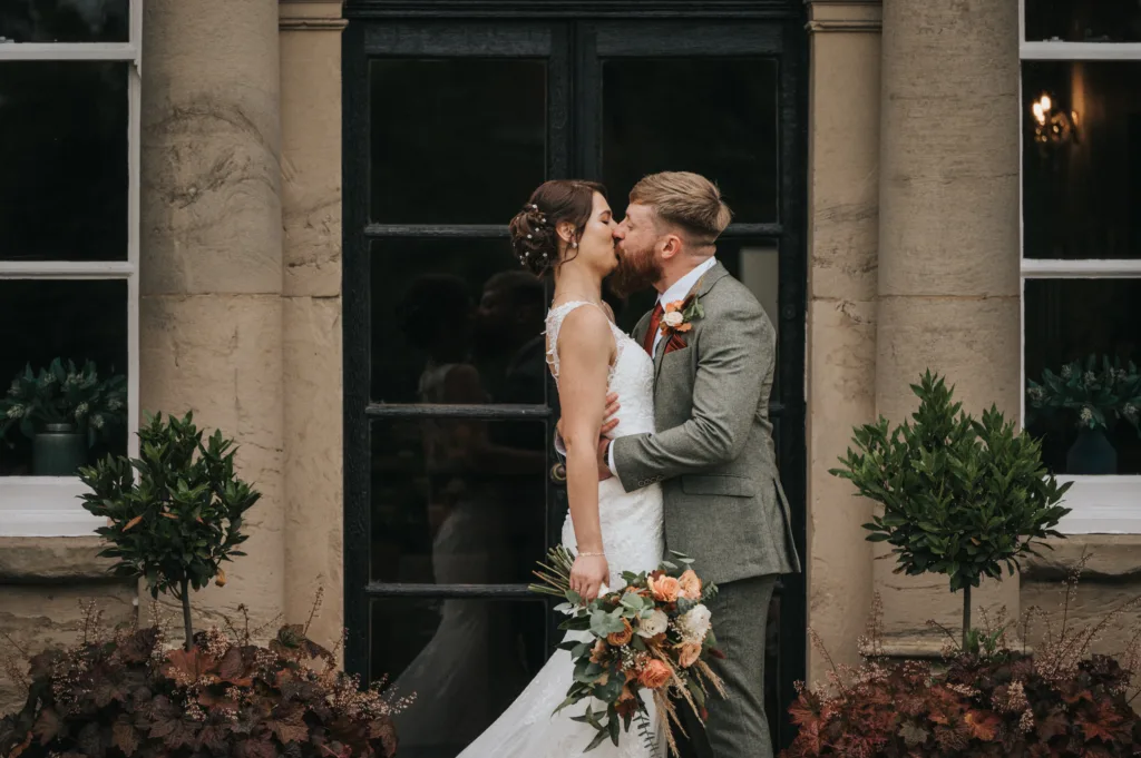A bride and groom kiss outside a building with large windows and pillars. The bride wears a white lace gown and holds a bouquet of orange and white flowers; the groom wears a light grey suit. Two green potted plants frame them on either side. © Aimee Lince Photography, Lincolnshire photographer