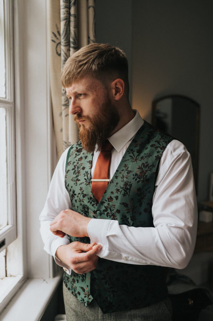 A bearded man stands by a window, adjusting his shirt cuff. He wears a white shirt, a rust-coloured tie, and a dark green floral-patterned waistcoat. Soft daylight enters the room, highlighting his focused expression. A mirror and beige curtains are visible in the background. © Aimee Lince Photography, Lincolnshire photographer