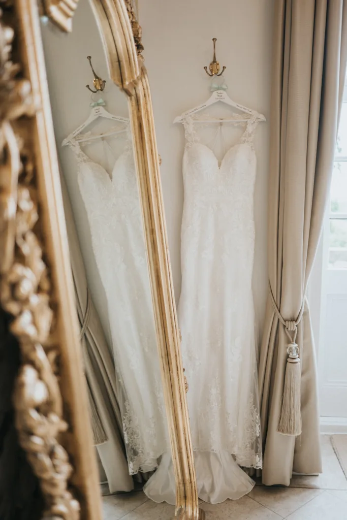 A white lace wedding dress with a fitted bodice and cap sleeves hangs on a hook, reflected in an ornate gold mirror. Beige curtains with a tassel tie-back frame a nearby window, creating a soft, elegant atmosphere. © Aimee Lince Photography, Lincolnshire photographer