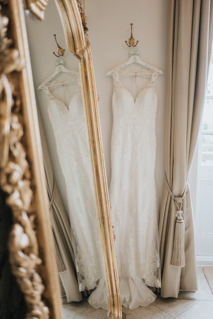 A white lace wedding dress with a fitted bodice and cap sleeves hangs on a hook, reflected in an ornate gold mirror. Beige curtains with a tassel tie-back frame a nearby window, creating a soft, elegant atmosphere. © Aimee Lince Photography, Lincolnshire photographer