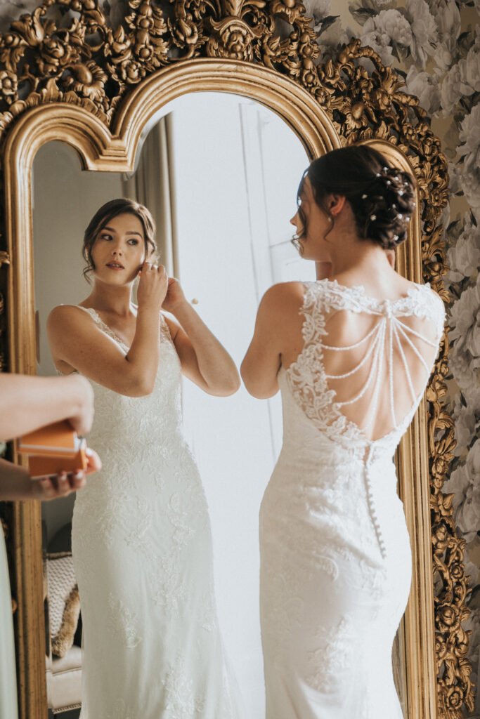 A bride in a white lace gown fastens an earring while standing before an ornate gold mirror. The dress features decorative back details with lace and bead strands. Her hair is styled in an elegant updo, and she looks at her reflection. © Aimee Lince Photography, Lincolnshire photographer