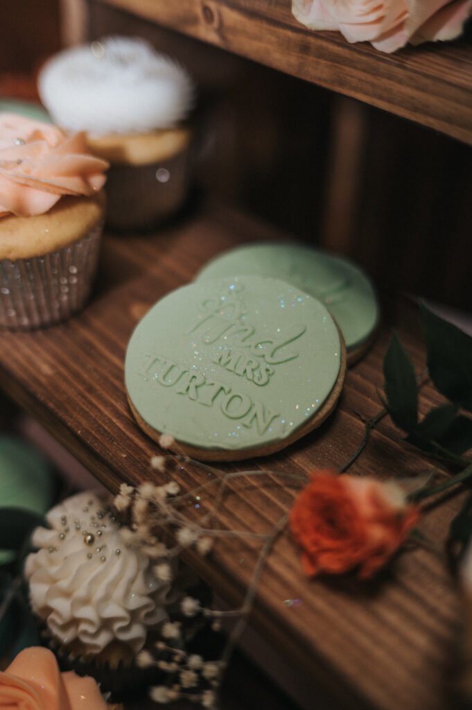 A close-up of mint-green round biscuits with the embossed text "3rd Years Turston," displayed on a rustic wooden shelf near peach and white iced cupcakes and orange flowers. The scene is elegant, with a celebratory and floral theme. © Aimee Lince Photography, Lincolnshire photographer
