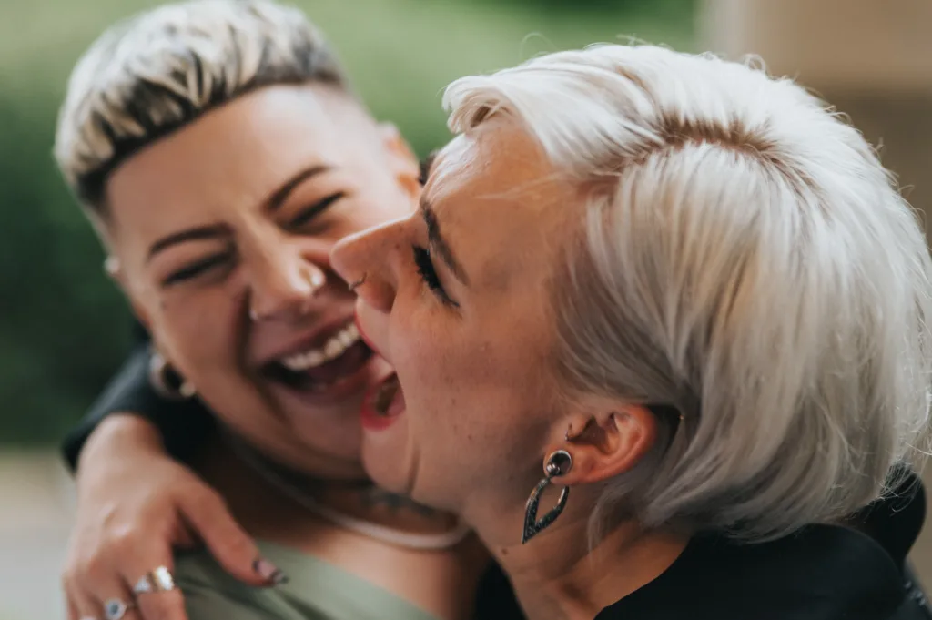 Two people with short, light-coloured hair laugh joyfully together. One has platinum blonde hair and black earrings, while the other has a shaved undercut. Their faces are close and one has their arm affectionately around the other, both radiating happiness. © Aimee Lince Photography, Lincolnshire photographer