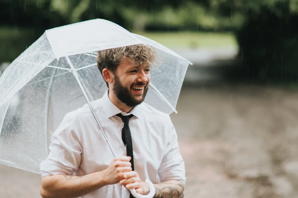 A smiling man with curly hair and a beard holds a clear umbrella while standing in the rain. He wears a white shirt with rolled sleeves and a black tie, with trees and a wet path visible in the blurred background. © Aimee Lince Photography, Lincolnshire photographer