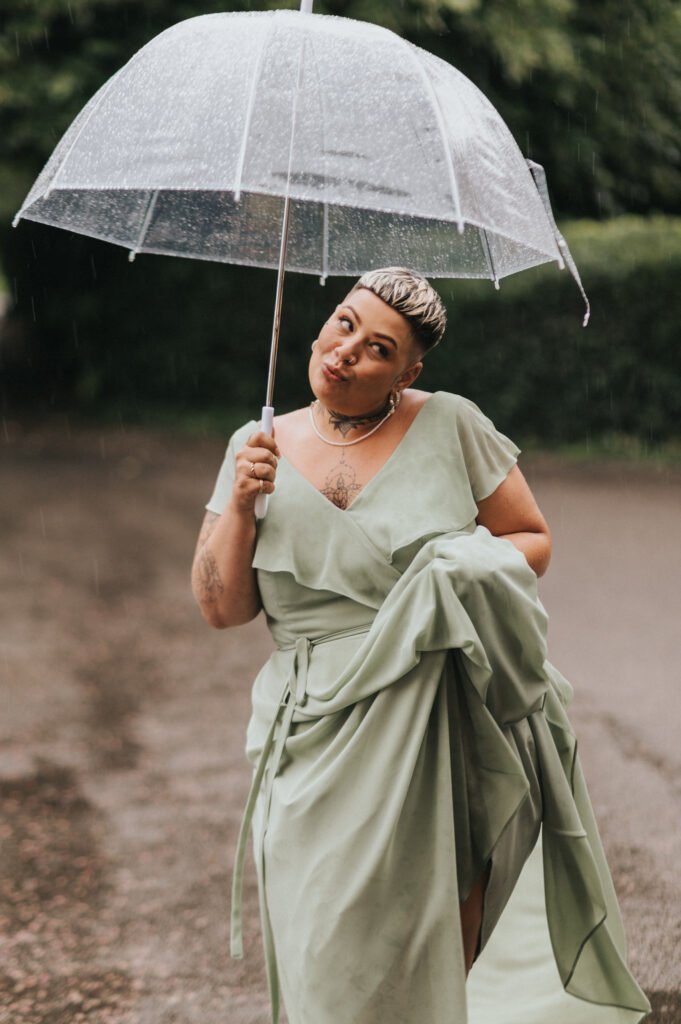 A person in a light green dress stands on a wet path, holding up the skirt with one hand and a clear umbrella with the other. Raindrops fall around them. They have short, slicked-back hair and wear a necklace, posing with a playful expression outdoors. © Aimee Lince Photography, Lincolnshire photographer