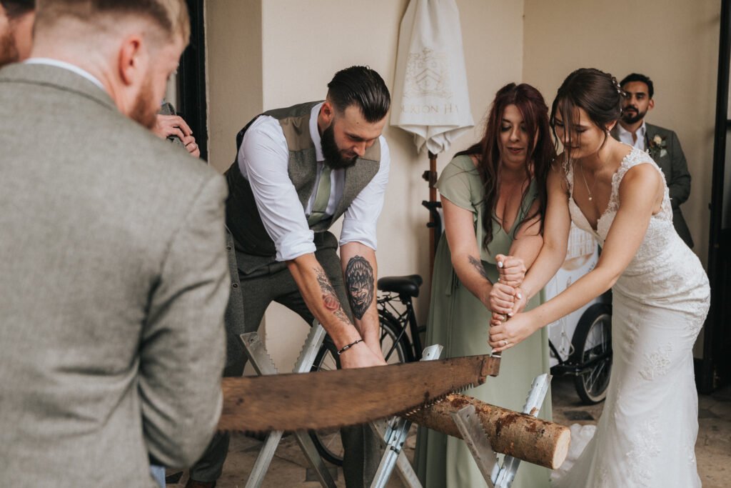 A bride in a white lace dress and a woman in green use a large saw to cut a log, while a man in a grey waistcoat helps guide the blade. Onlookers in suits and dresses watch the activity in a light-coloured indoor setting. © Aimee Lince Photography, Lincolnshire photographer