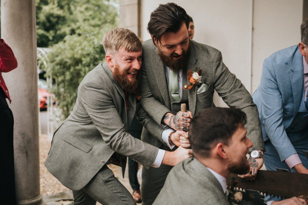 Three men in suits, two with beards and tattoos, are laughing and sawing a wooden log together at an outdoor event. One man in a blue suit stands partly visible on the right. The scene appears joyful and lively. © Aimee Lince Photography, Lincolnshire photographer