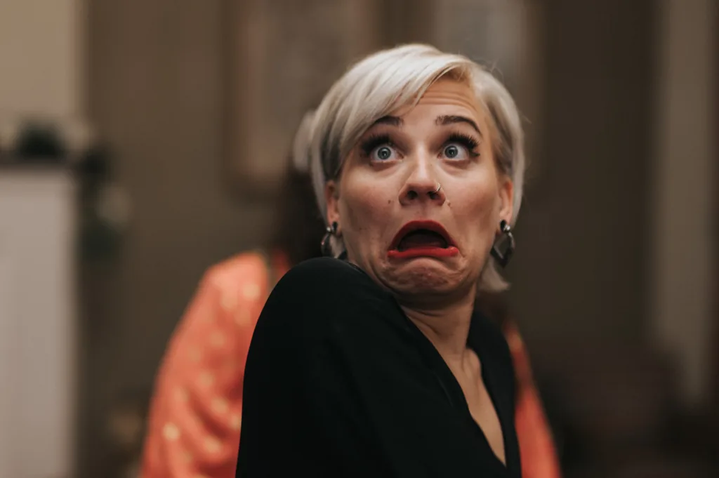 A woman with short platinum blonde hair and dark earrings looks surprised or shocked, eyes wide and mouth open in an exaggerated expression. She wears a black top and stands indoors with blurred background elements. © Aimee Lince Photography, Lincolnshire photographer