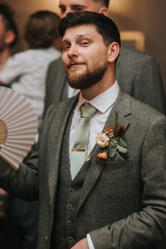 A bearded man in a grey suit with a floral buttonhole and pale green tie smirks at the camera, holding a white folding fan. People are blurred in the background, giving the scene a lively, celebratory feel. © Aimee Lince Photography, Lincolnshire photographer