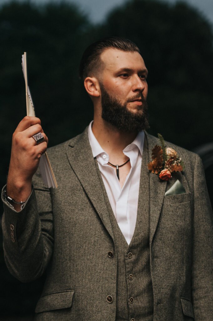 A bearded man in a grey suit and open-collar white shirt stands outdoors, holding up a stack of papers—perhaps vows—for a wedding photographer to capture. He wears rings, a necklace, and a buttonhole, with blurred greenery in the background. © Aimee Lince Photography, Lincolnshire photographer