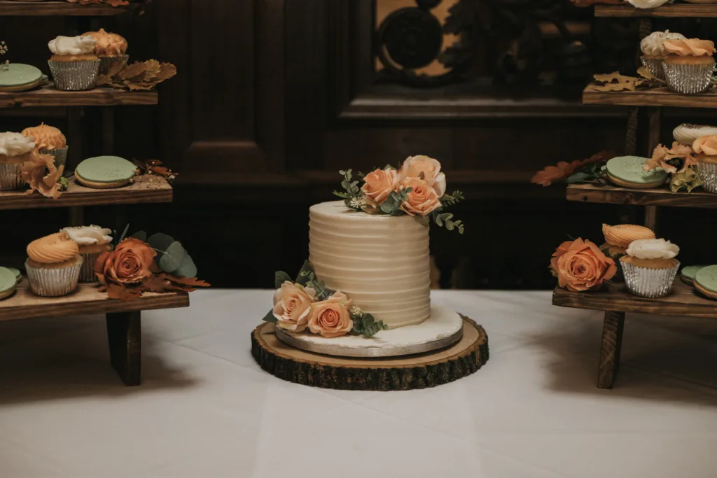 A small, white tiered cake with textured icing is decorated with peach roses and greenery, set on a wooden slab. Surrounding it are rustic wooden stands holding cupcakes and macarons, also adorned with peach flowers. The background features dark wood panelling. © Aimee Lince Photography, Lincolnshire photographer