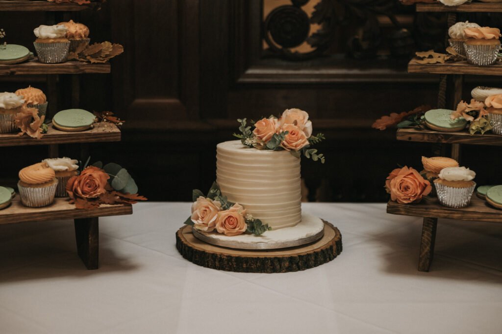 A small, white tiered cake with textured icing is decorated with peach roses and greenery, set on a wooden slab. Surrounding it are rustic wooden stands holding cupcakes and macarons, also adorned with peach flowers. The background features dark wood panelling. © Aimee Lince Photography, Lincolnshire photographer