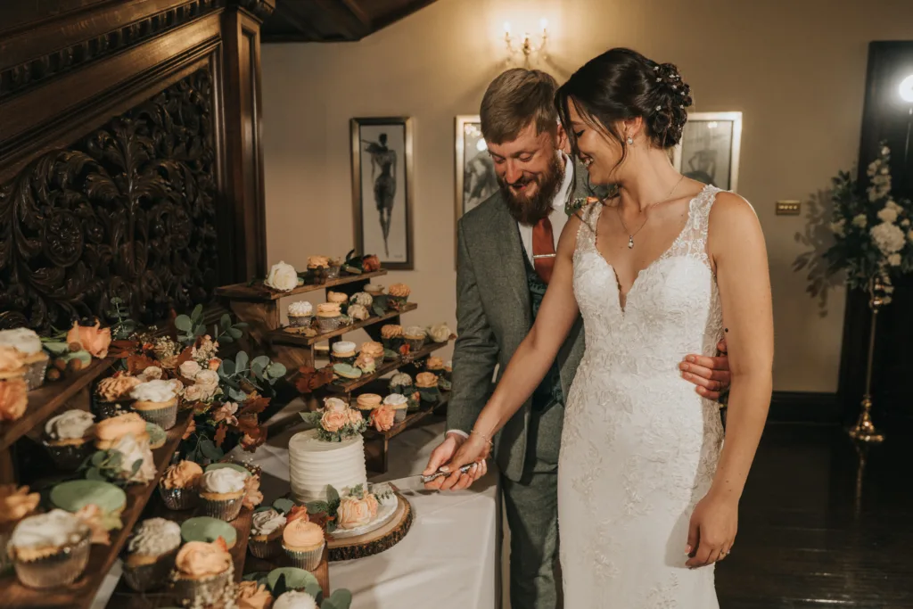 A bride in a white lace gown and a groom in a grey suit smile as they cut a small white wedding cake together. They are surrounded by elegant displays of assorted cupcakes with peach and cream icing in a warmly lit room. © Aimee Lince Photography, Lincolnshire photographer