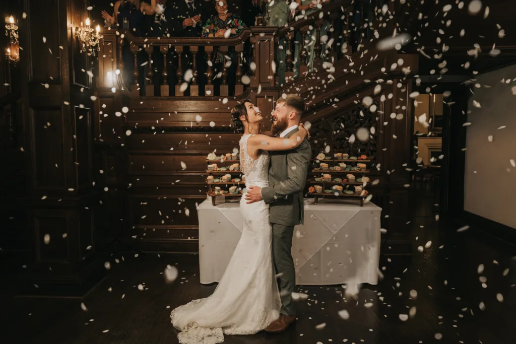 A bride and groom in wedding attire embrace and smile at each other while dancing indoors beneath a large wooden staircase at Bourton Hall. They are surrounded by falling flower petals, with a grand wooden staircase and guests in the background, creating a romantic and festive atmosphere. © Aimee Lince Photography, Lincolnshire photographer