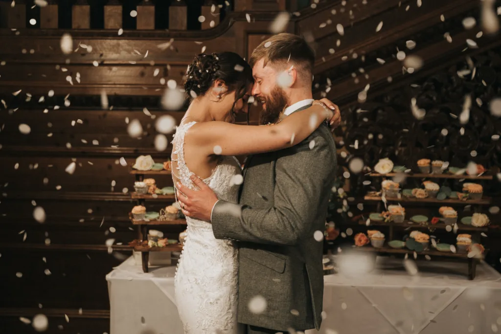A bride in a white lace dress and a groom in a grey suit embrace and smile during their first dance, surrounded by falling confetti. In the background, there is a table with a cupcake display and ornate wood panelling on the walls. © Aimee Lince Photography, Lincolnshire photographer