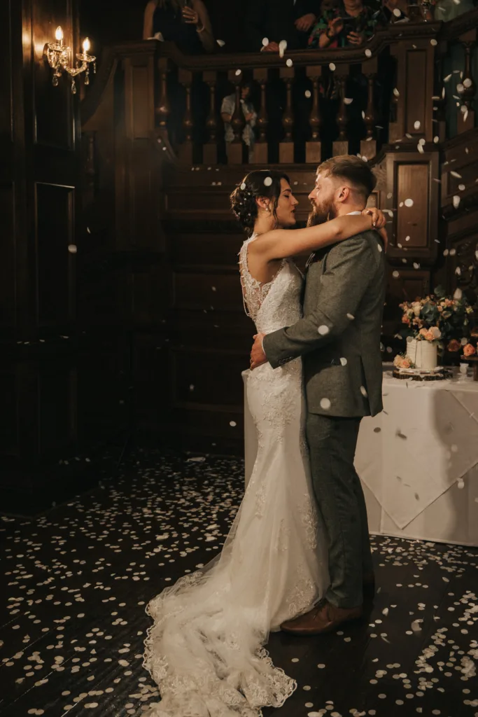 A bride in a white lace wedding dress and a groom in a grey suit embrace and dance together on a dark wooden floor covered in white confetti. Behind them are wooden stairs, onlookers above, and a table decorated with flowers and a cake. © Aimee Lince Photography, Lincolnshire photographer