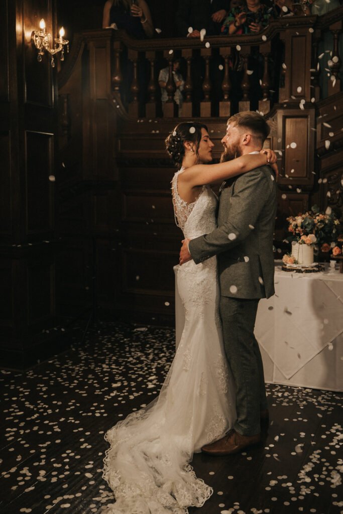 A bride in a white lace wedding dress and a groom in a grey suit embrace and dance together on a dark wooden floor covered in white confetti. Behind them are wooden stairs, onlookers above, and a table decorated with flowers and a cake. © Aimee Lince Photography, Lincolnshire photographer