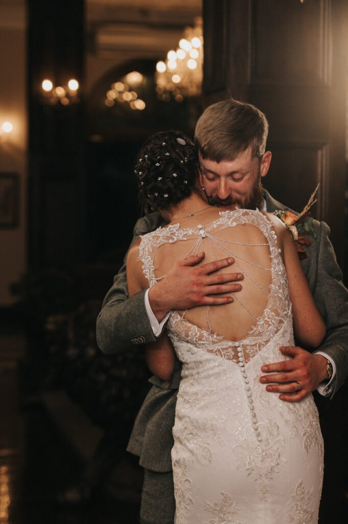 A man in a grey suit embraces a woman in a white lace wedding dress with an open back and delicate bead details. The couple shares an intimate hug indoors, with warm lighting and soft chandeliers glowing in the background. © Aimee Lince Photography, Lincolnshire photographer