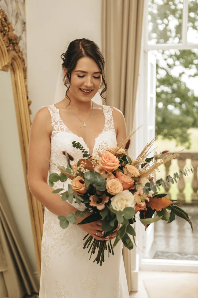 A smiling bride in a white lace wedding dress holds a large bouquet of peach and cream roses, greenery, and rustic foliage. She stands indoors by a tall window with an ornate gold mirror and beige curtains in the background. © Aimee Lince Photography, Lincolnshire photographer