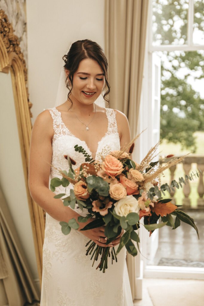 A smiling bride in a white lace wedding dress holds a large bouquet of peach and cream roses, greenery, and rustic foliage. She stands indoors by a tall window with an ornate gold mirror and beige curtains in the background. © Aimee Lince Photography, Lincolnshire photographer
