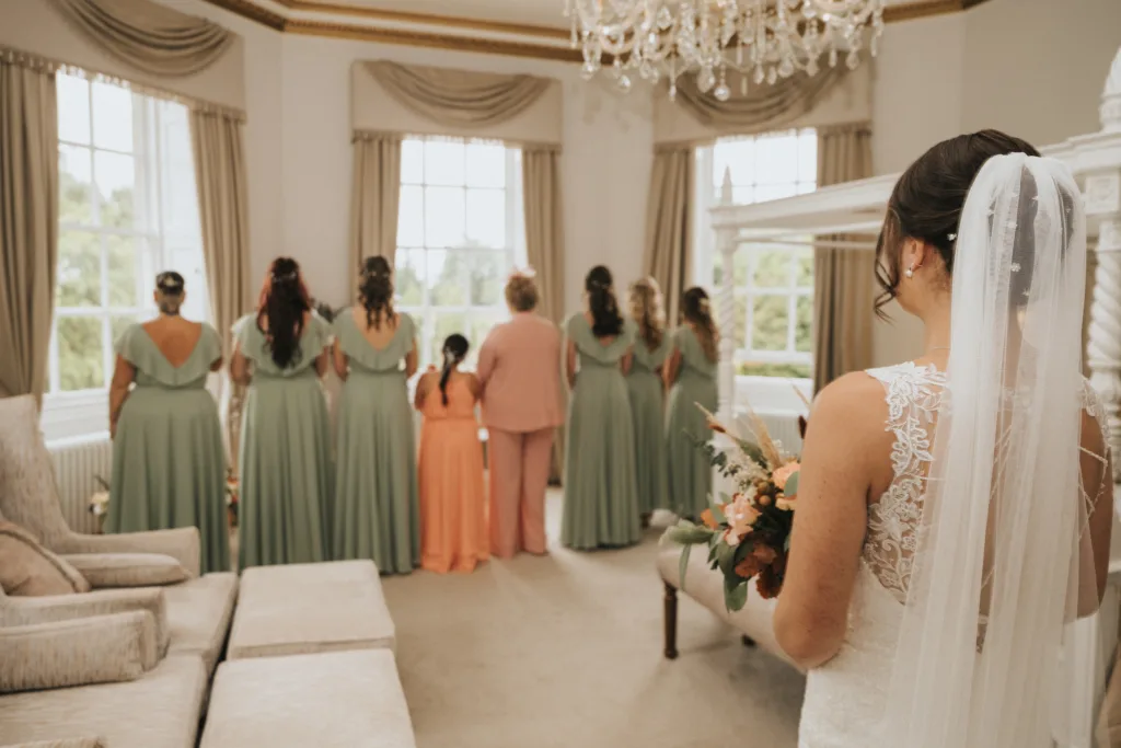A bride in a lace wedding dress and veil holds a bouquet, standing in a bright, elegant room. Ahead, seven women in sage green and peach dresses and one woman in a peach suit stand with their backs to her, looking out large windows with beige curtains. © Aimee Lince Photography, Lincolnshire photographer